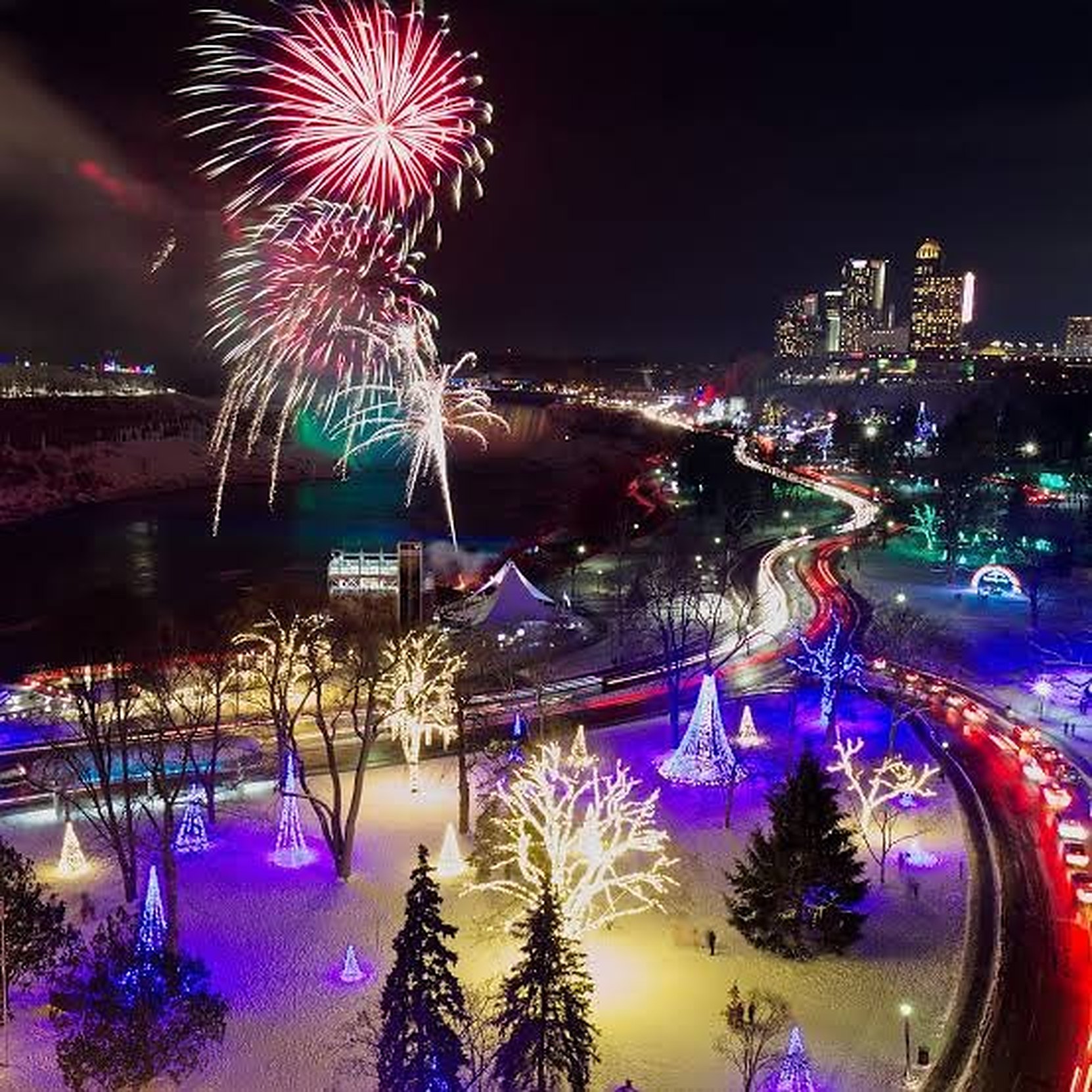 Fireworks bursting above snow-covered gardens hung with winter festival lights, with light trails along the road beyond